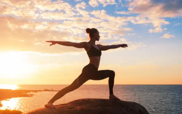 Yoga anksiyete için düzenli pratik — gün batımında kayalıkta savaşçı II pozunda kadın