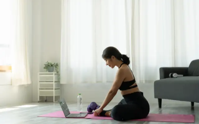 Evde yoga yapan bir kadının fotoğrafı. Matına oturmuş videodan yoga dersi takip ediyor.